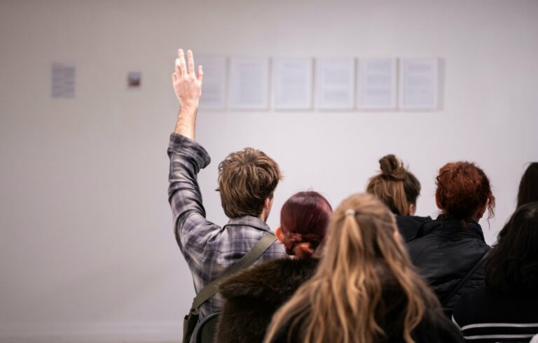 Dans une salle vue de dos, un élève lève la main pendant l'intervention d'un conférencier