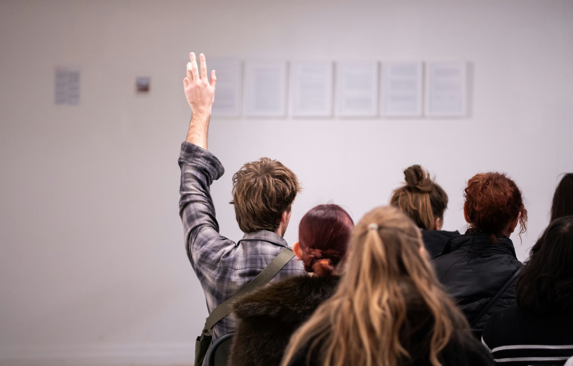 Dans une salle vue de dos, un élève lève la main pendant l'intervention d'un conférencier